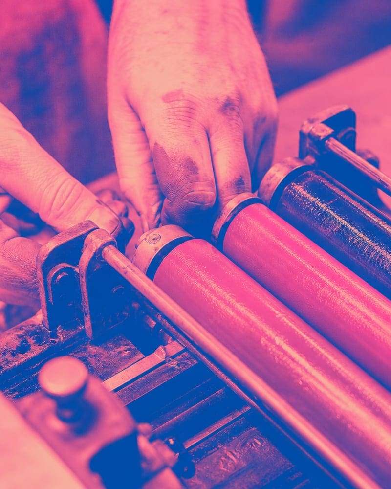 Hands adjusting ink rollers on a Heidelberg windmill press, fluoro pink and blue visible on the rollers.