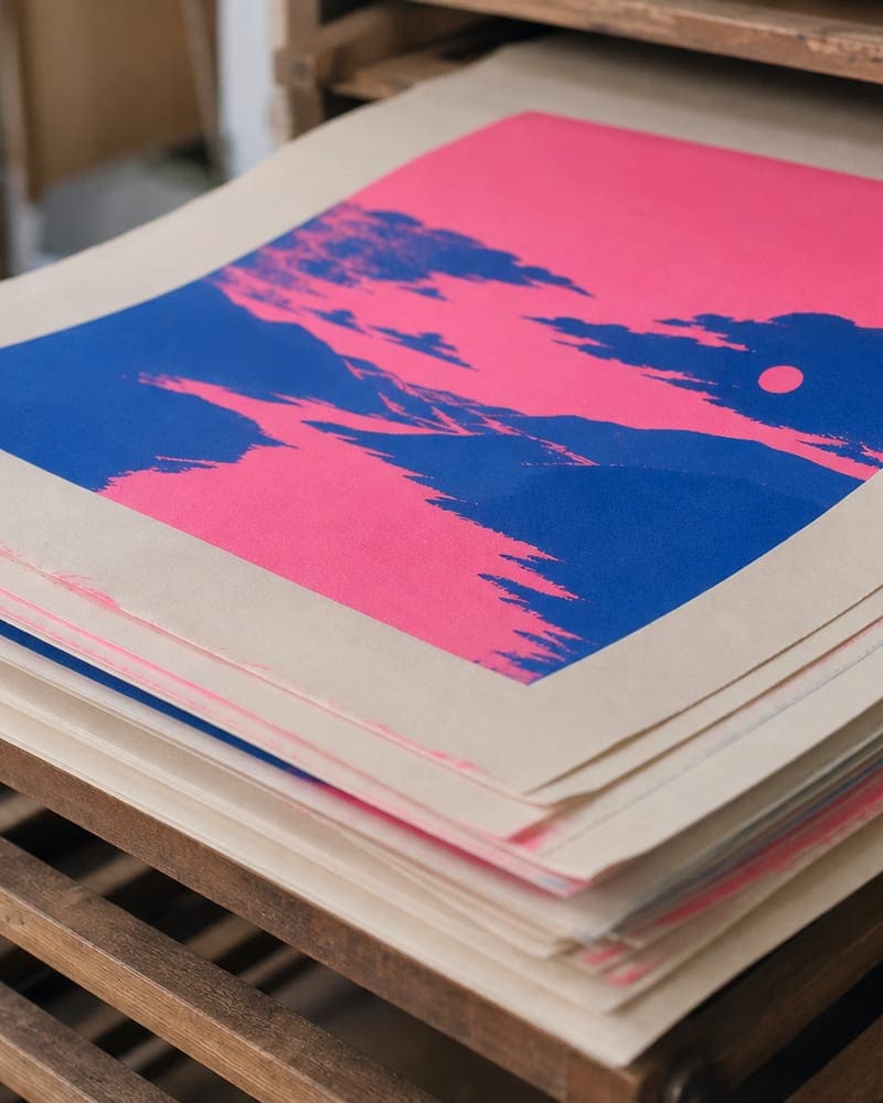 Stack of risograph prints drying on a wooden rack, edges slightly feathered with ink bleed.